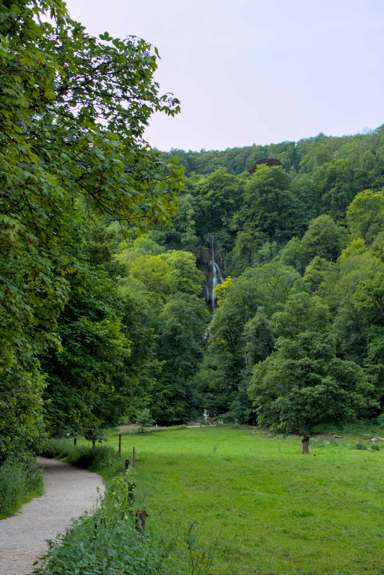 Ein Tal in einer bergigen Gegend mit einem hohen Wasserfall am Ende, darum viele Bäume und davor ein Wanderweg neben einer grünen Wiese